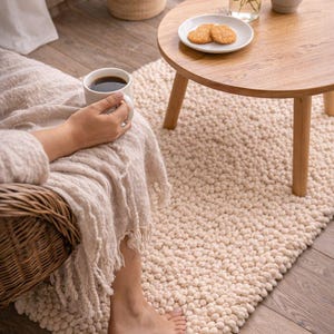 May include: A cozy outdoor scene featuring a small wooden table with a plate of cookies, a white mug of coffee, and a cream-colored rug. A wicker chair and potted plants add to the relaxing atmosphere. The rug is made of a soft, textured material.