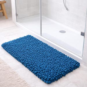May include: A rectangular, textured, blue bath mat. The mat is made of a looped material and is placed on a tiled floor in front of a shower. The mat's dimensions are approximately 30 inches long and 20 inches wide.