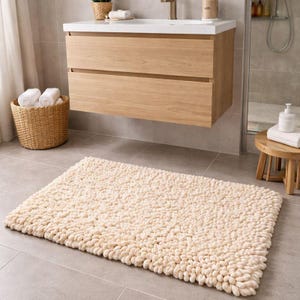 May include: Cream-colored, rectangular bath mat with a textured, looped design. The mat is placed on a gray tiled floor in a bathroom setting. A wooden vanity with a white countertop and a woven basket are also visible.