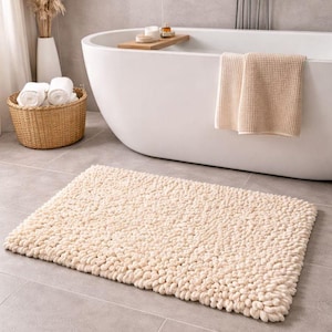 Op de afbeelding: Een crèmekleurige, rechthoekige badmat met een getextureerd, gelust oppervlak. De mat ligt op een grijze tegelvloer voor een witte badkuip. Een gevlochten mand met opgerolde witte handdoeken en een witte vaas staan op de achtergrond.