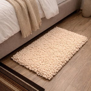 May include: A cream-colored, rectangular shag rug with a dense, textured surface. The rug is placed on a wooden floor near a bed with a beige blanket. A smaller brown and beige patterned rug is also visible.