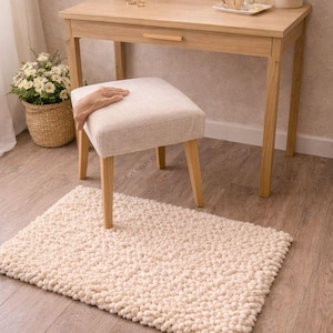 May include: A light wood vanity table with a matching stool and a cream-colored, textured rug. The table has a round mirror, a drawer, and various decorative items. A woven basket with flowers is on the left.