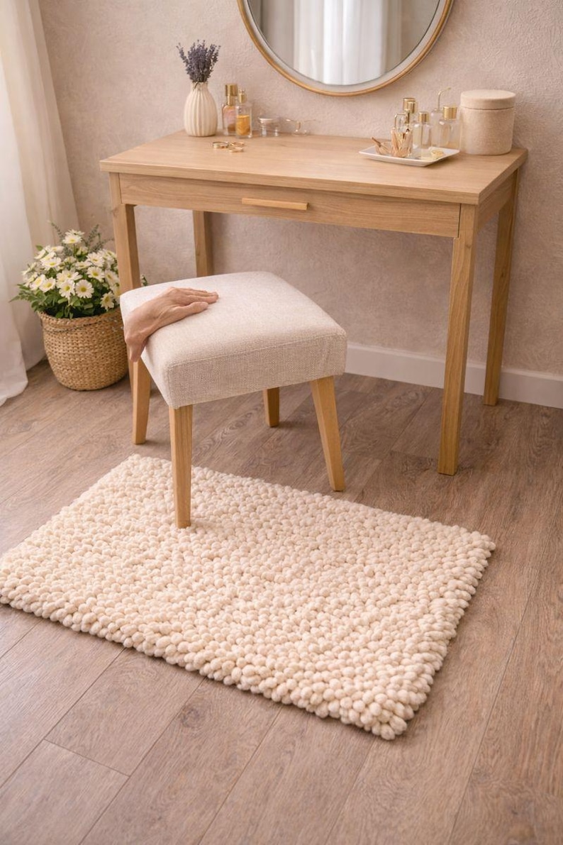 May include: A light wood vanity table with a matching stool and a cream-colored, textured rug. The table has a round mirror, a drawer, and various decorative items. A woven basket with flowers is on the left.