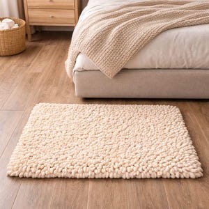 May include: A rectangular, cream-colored, textured rug sits on a hardwood floor. The rug has a thick, looped pile, and is placed in front of a bed with a beige blanket. A wooden nightstand and a wicker basket are also visible.