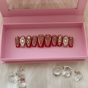 May include: A set of ten false nails in a pink box. The nails are a light brown colour with a red tip and gold and white embellishments.