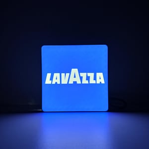 May include: A square, illuminated sign with a bright blue face. The white text "LAVAZZA" is centered on the sign. The sign is lit against a dark blue background, creating a glowing effect.