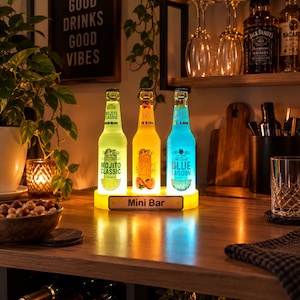 Può includere: Un espositore per minibar luminoso con tre bottiglie di vetro illuminate etichettate Mojito Classic, Sex on the Beach e Blue Lagoon. Le bottiglie brillano di verde, arancione e blu, poggiate su una base gialla con la scritta Mini Bar stampata.