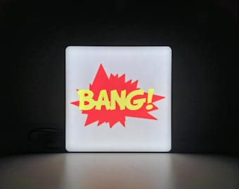 May include: A square, illuminated sign with the word "BANG!" in yellow, set against a red burst graphic. The sign has a white border and is lit from within. The background is dark.