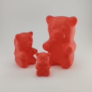 Könnte beinhalten: Drei rote Gummibärchen-Figuren in verschiedenen Größen sind abgebildet. Der größte Bär steht aufrecht, die anderen beiden sind kleiner. Die Bären haben eine glatte, glänzende Oberfläche und sind in einem leuchtenden Rot gehalten. Auf weißem Hintergrund.