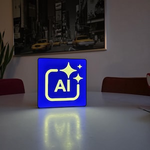 Könnte beinhalten: Ein blaues quadratisches Licht mit den weißen Buchstaben "AI", umgeben von einem weißen Rand und zwei gelben Sternen. Das Licht steht auf einem weißen Tisch. Im Hintergrund hängt ein schwarz-weißes Stadtbildgemälde.