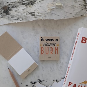 May include: A small, brown notepad with the text "it was a pleasure to burn" printed on the cover. The notepad is sitting on a white surface with a pencil and a book titled "Fahrenheit 451" by Ray Bradbury.