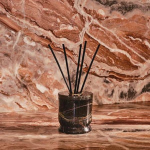 Puede incluir: Un difusor de caña de mármol negro con cañas negras. El difusor cilíndrico tiene detalles dorados y un diseño estampado. El fondo es una superficie de mármol marrón rojizo.