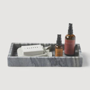 Puede incluir: Una bandeja rectangular gris con aspecto de mármol contiene una pastilla de jabón, un paño y dos botellas con pulverizador. El jabón es blanco con la palabra "CLOVER" impresa. Las botellas son de vidrio ámbar con pulverizadores negros.