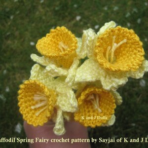 May include: A bouquet of crocheted daffodils in yellow and white. The flowers are arranged in a hand, with the text "K and J Dolls" and "Daffodil Spring Fairy crochet pattern by Sayjai of K and J Dolls" visible at the bottom of the image.