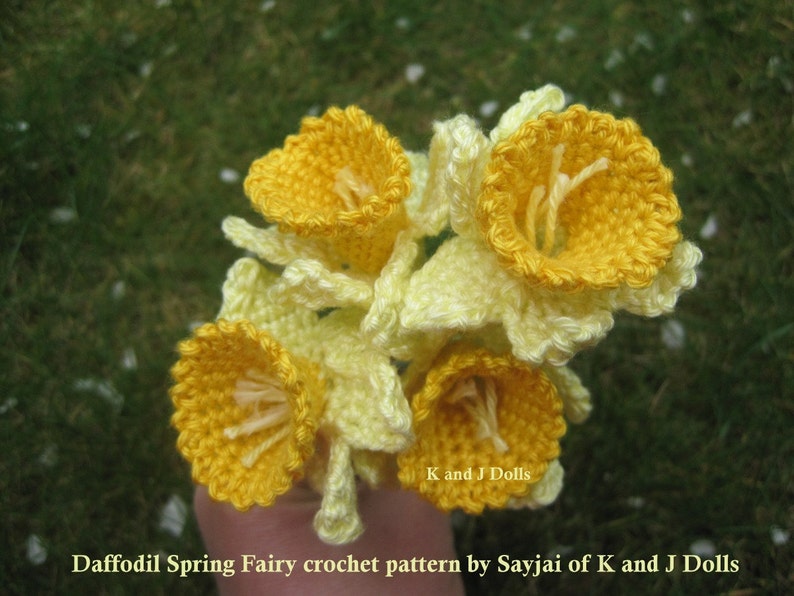 May include: A bouquet of crocheted daffodils in yellow and white. The flowers are arranged in a hand, with the text "K and J Dolls" and "Daffodil Spring Fairy crochet pattern by Sayjai of K and J Dolls" visible at the bottom of the image.