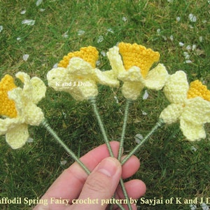 May include: Five crocheted daffodil flowers with yellow centers and white petals. The flowers are held in a hand against a green grassy background.