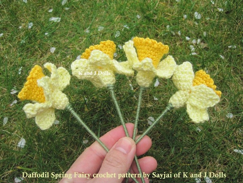 May include: Five crocheted daffodil flowers with yellow centers and white petals. The flowers are held in a hand against a green grassy background.