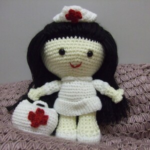 May include: A crocheted doll dressed as a nurse, wearing a white dress, a red cross hat, and carrying a small red cross bag. The doll has black hair and a smiling face.