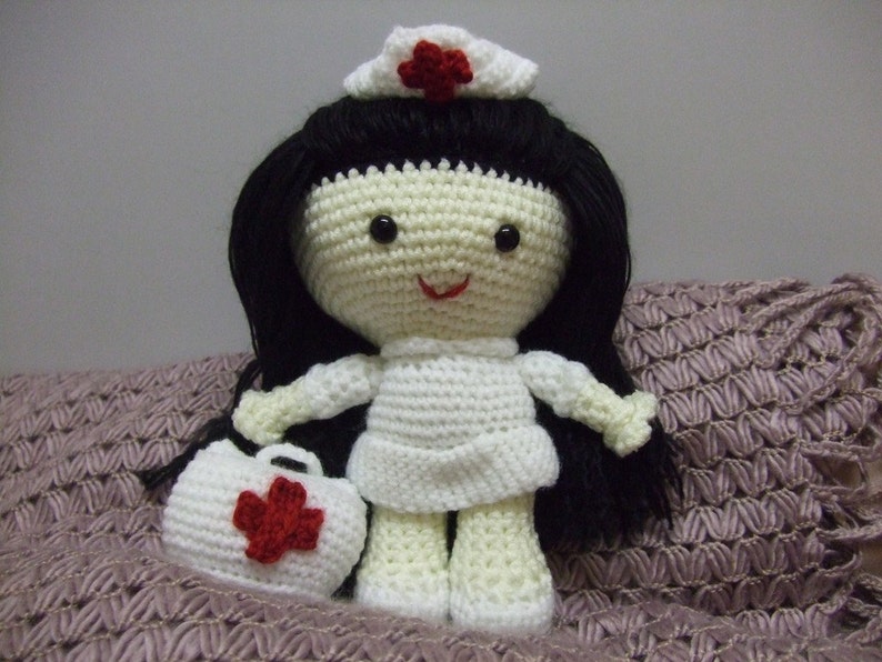 May include: A crocheted doll dressed as a nurse, wearing a white dress, a red cross hat, and carrying a small red cross bag. The doll has black hair and a smiling face.