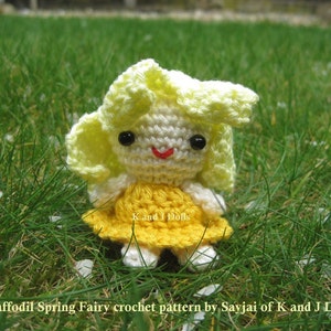 May include: A crocheted fairy doll with yellow hair and a yellow dress. The doll is sitting in a bed of green grass with white flower petals scattered around it. The doll's eyes are black and her mouth is a red smile. The doll is labeled "Daffodil Spring Fairy crochet pattern by Sayjai of K and J Dolls".