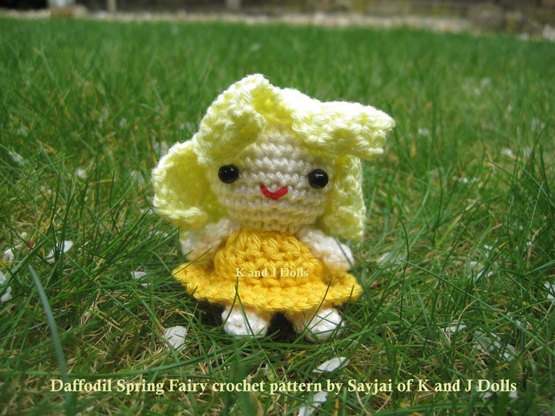 May include: A crocheted fairy doll with yellow hair and a yellow dress. The doll is sitting in a bed of green grass with white flower petals scattered around it. The doll's eyes are black and her mouth is a red smile. The doll is labeled "Daffodil Spring Fairy crochet pattern by Sayjai of K and J Dolls".