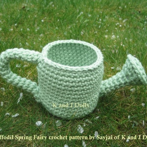 May include: A light green crocheted watering can with a handle. The watering can is sitting on a bed of green grass with white flower petals scattered around it.