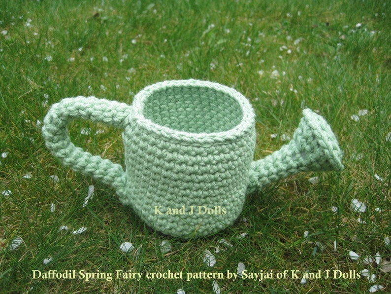 May include: A light green crocheted watering can with a handle. The watering can is sitting on a bed of green grass with white flower petals scattered around it.