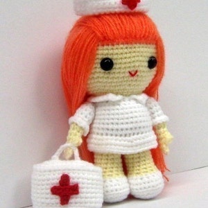 May include: A crocheted doll dressed as a nurse, wearing a white dress, a white hat with a red cross, and holding a white bag with a red cross. The doll has orange hair and a smiling face.