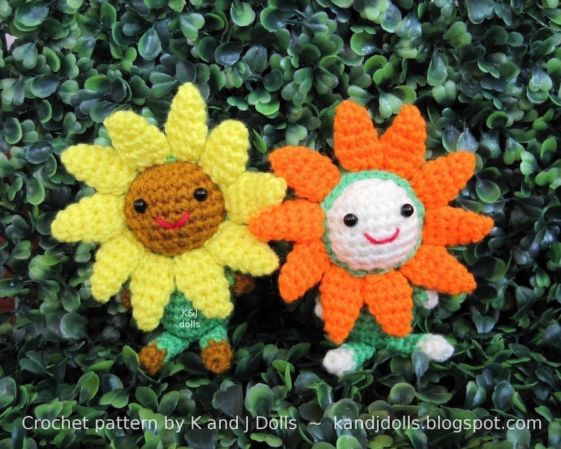 May include: Two crocheted sunflower dolls with smiling faces. One is yellow and brown, the other is orange and white. Both have green and brown accents. The dolls are set against a backdrop of green foliage. The text "Crochet pattern by K and J Dolls" is visible.