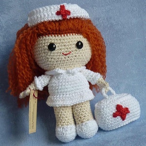 May include: Crocheted doll dressed as a nurse with a red cross on her hat and a white crocheted bag with a red cross. The doll has red hair and brown eyes.