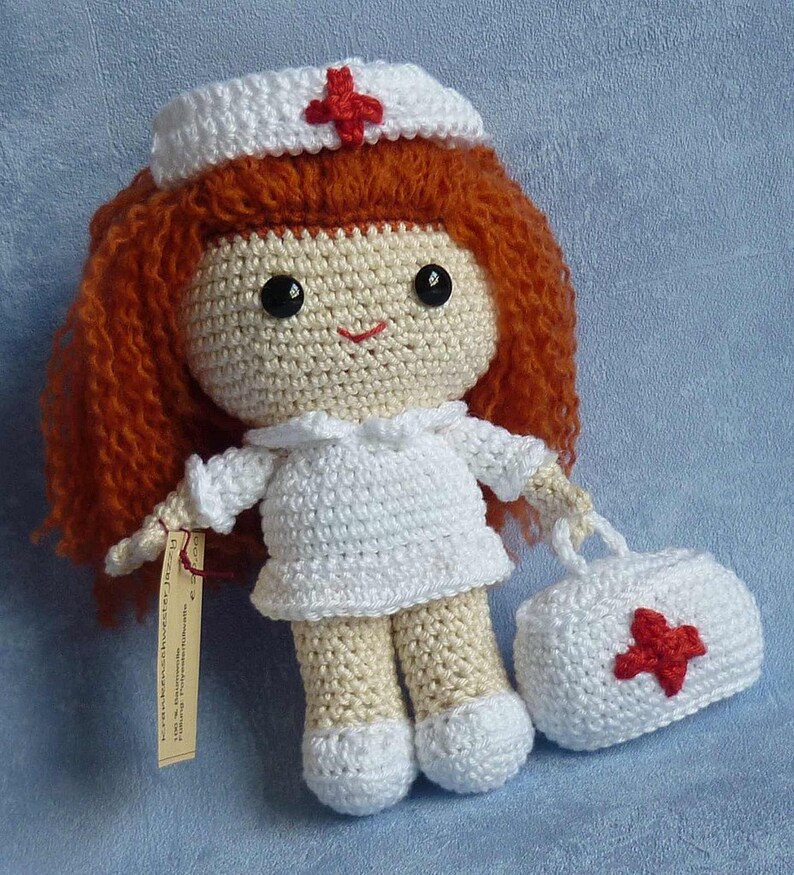 May include: Crocheted doll dressed as a nurse with a red cross on her hat and a white crocheted bag with a red cross. The doll has red hair and brown eyes.
