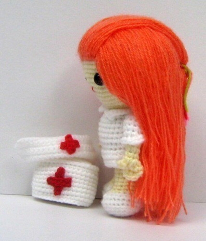 May include: A crocheted doll dressed as a nurse with a white dress, red crosses on her hat, and long orange hair.