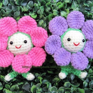May include: Two handmade crochet flower dolls. One doll has pink petals, the other has purple petals. Both dolls have white faces with black eyes and red smiles. The dolls have green stems and feet. The dolls are set against a background of green leaves.