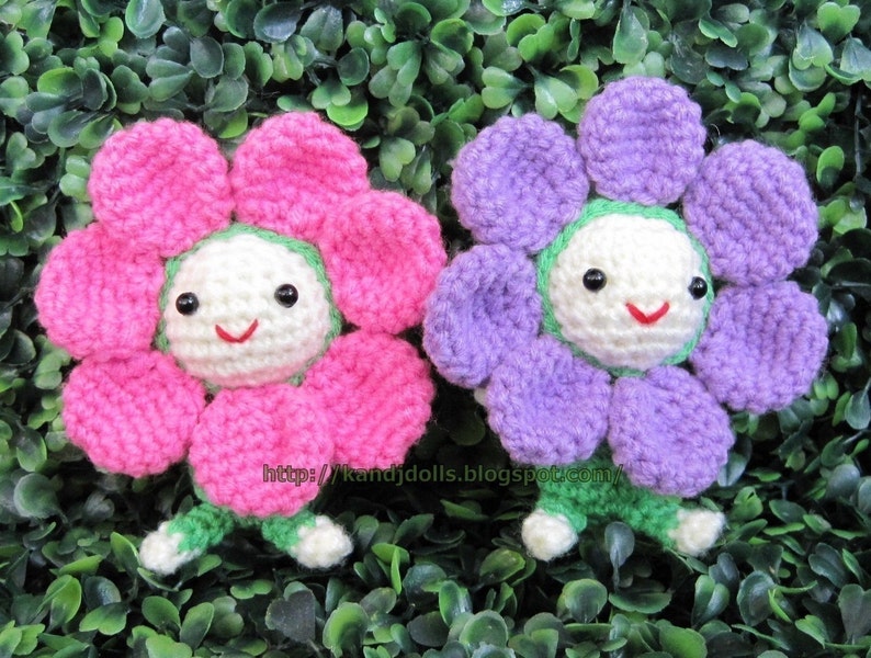 May include: Two handmade crochet flower dolls. One doll has pink petals, the other has purple petals. Both dolls have white faces with black eyes and red smiles. The dolls have green stems and feet. The dolls are set against a background of green leaves.