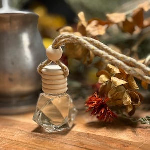 May include: A clear, geometric-shaped glass bottle with a wooden top and a beige rope for hanging. The bottle contains a light-colored liquid. The background features dried flowers and foliage, creating a natural aesthetic.