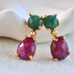 May include: A pair of gold-tone drop earrings featuring a faceted round green stone at the top, a small white pearl accent in the center, and a faceted pear-shaped deep pink stone at the bottom, all set in a gold-colored metal frame.