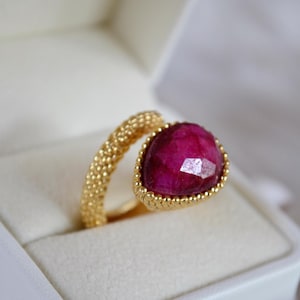 Pode incluir: Um anel de ouro com uma grande pedra preciosa facetada de cor rubi. A banda do anel tem um design texturizado e com contas. A pedra preciosa é engastada numa moldura de ouro com uma borda de contas. O anel é apresentado numa caixa de joias branca.