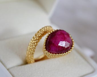 Può includere: Un anello dorato con una fascia testurizzata a perline e una singola pietra preziosa rosa scuro, sfaccettata a forma di pera e incastonata in una montatura dorata. L'anello è mostrato in una scatola bianca, evidenziando il contrasto tra oro e rosa.