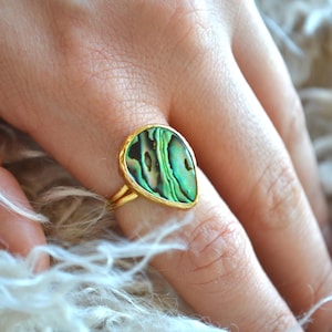 Peut inclure: Une bague dorée ornée d'une coquille d'ormeau en forme de goutte d'eau, aux motifs irisés verts et noirs. Le sertissage de la bague présente un aspect martelé. La bague est sur un doigt.