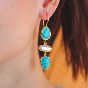 May include: Gold-tone drop earrings featuring turquoise teardrop and rectangular white stone accents. The earrings have a gold frame around each stone and a hook closure. The earrings are shown on a person's ear.