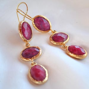 May include: A pair of gold-toned drop earrings featuring three faceted, oval-shaped deep magenta stones set in textured metal bezels. Each earring hangs from a simple gold-toned wire hook, showcasing a vertical arrangement of the stones.