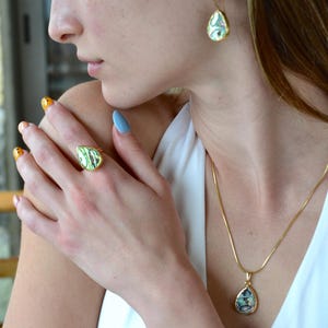 Könnte beinhalten: Ein goldfarbenes Schmuckset bestehend aus einem tropfenförmigen Ring, einem passenden tropfenförmigen Anhänger an einer dünnen Goldkette und einem einzelnen tropfenförmigen Ohrring. Jedes Stück hat eine irisierende Abalone-Muschel-Einlage.