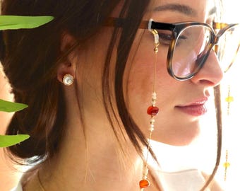 May include: A beaded eyeglass chain featuring small white beads, gold-toned spacers, and larger smooth orange-red stones. The chain attaches to the temples of brown tortoiseshell-patterned eyeglasses with clear silicone loops and gold-toned metal hardware.