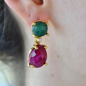 Puede incluir: Un pendiente colgante de oro con una piedra verde redonda facetada en la parte superior, una pequeña perla blanca en el centro y una piedra magenta ovalada facetada en la parte inferior, todo montado en un marco de metal dorado con garras.