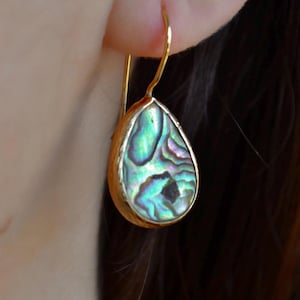 Op de afbeelding: Een druppelvormige oorbel met een goudkleurig montuur en een glinsterende abalone schelpinleg. De schelp toont iriserende blauwe, groene en paarse tinten. De oorbel hangt aan een goudkleurige haak.