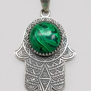 May include: A silver-toned metal Hamsa pendant featuring an intricate, embossed geometric pattern across its surface. A round, polished green malachite gemstone with dark swirling bands is set securely in the center of the palm area.