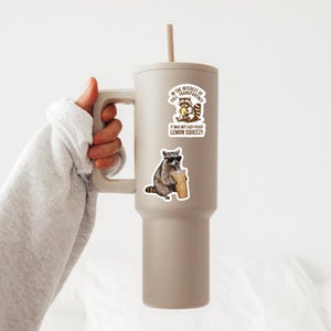 May include: A large, taupe-colored tumbler with a handle and a straw. The tumbler has two stickers featuring raccoons. One raccoon is eating a lemon, and the other is wearing sunglasses and drinking from a cup. Text on the stickers reads, "In the interest of full transparency" and "It was not easy peasy lemon squeezy."