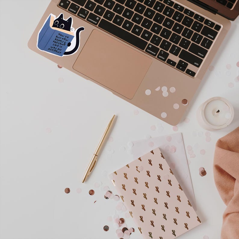 May include: A black cat peeking out from a blue book with the text "How to buy new books and pretend like it was an accident". The cat is on a laptop with a gold keyboard and a white background. There are pink and gold confetti scattered around the laptop.