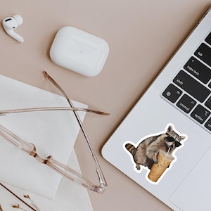 May include: A sticker of a raccoon wearing sunglasses and drinking iced coffee is affixed to a laptop. The raccoon is brown and gray with a striped tail. Other items include a pair of glasses, wireless earbuds, and a charging case.