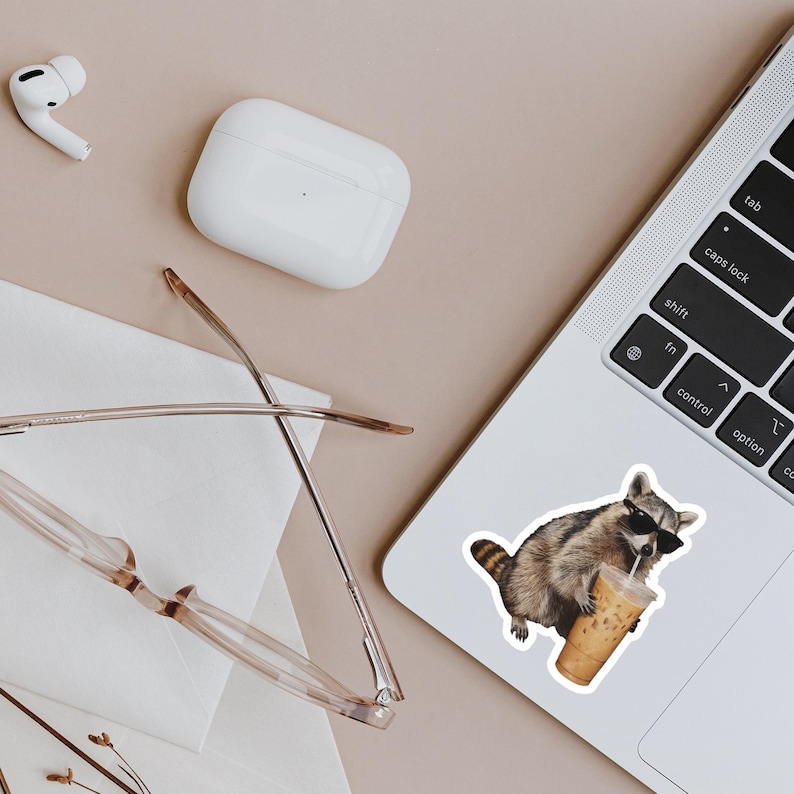 May include: A sticker of a raccoon wearing sunglasses and drinking iced coffee is affixed to a laptop. The raccoon is brown and gray with a striped tail. Other items include a pair of glasses, wireless earbuds, and a charging case.